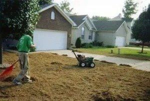 Man rakes mulch on a lawn in front of suburban houses, using a spreader.