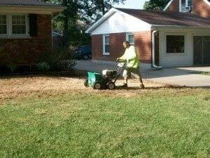 A person in a yellow vest spreads fertilizer on a lawn with a spreader, next to a driveway and houses.
