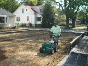 Man using a seed spreader on a lawn in front of a house; other workers are nearby.