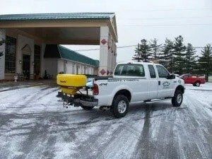 White pickup truck with a yellow salt spreader on a snowy driveway. The truck is parked in front of a building.