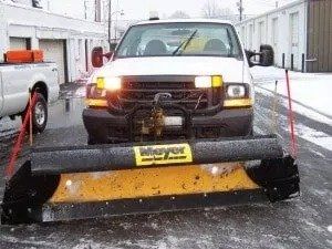 A white pickup truck with a yellow snowplow is parked on a snowy surface. The plow is lowered.