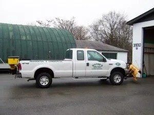 White pickup truck with a snowplow parked outside a building with a green arched roof. The truck has a salt spreader in the bed.