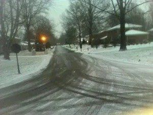 Snowy residential street with tire tracks, lined with leafless trees and houses. A street light glows in the distance.