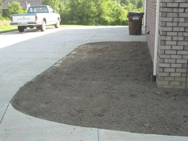 A driveway with a bare, prepared patch of soil next to a brick building. A white pickup truck is parked in the background.
