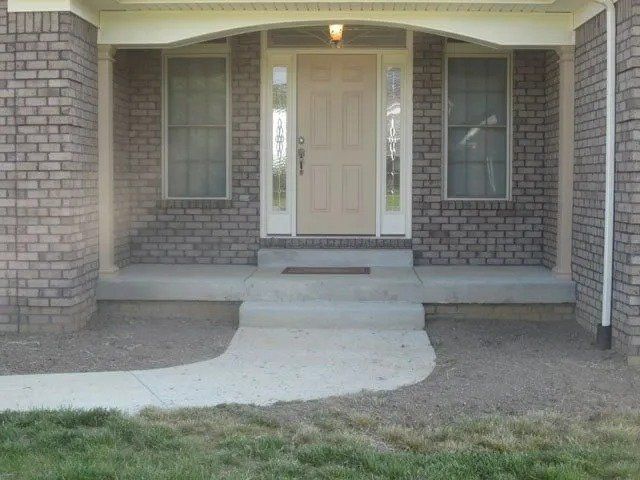 A brick home's front entrance with a concrete porch, steps, and a curved walkway; a light-colored door is flanked by windows.
