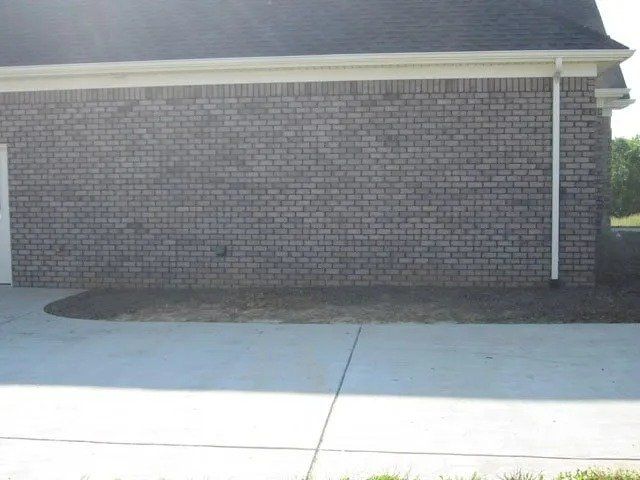 Brick exterior of a building with a concrete driveway and a strip of dirt along the foundation.