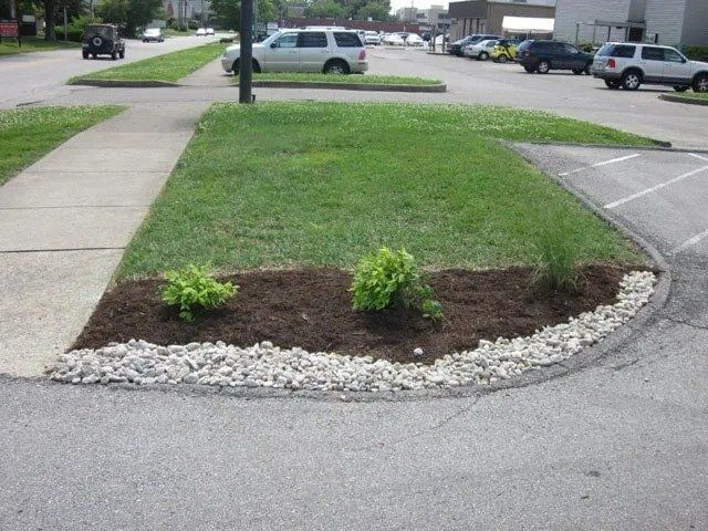 A landscaped island with green grass, mulch, and shrubs, bordered by a line of gray rocks and a paved road.