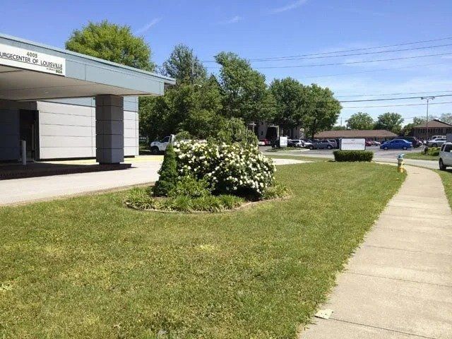 Exterior of a building with a small garden bed. A sidewalk leads past the building toward a parking area. Blue sky overhead.