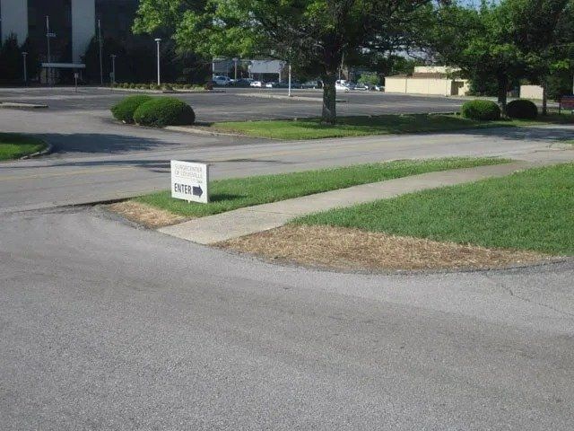 A sign on a patch of grass next to a road. A building and trees are in the background.