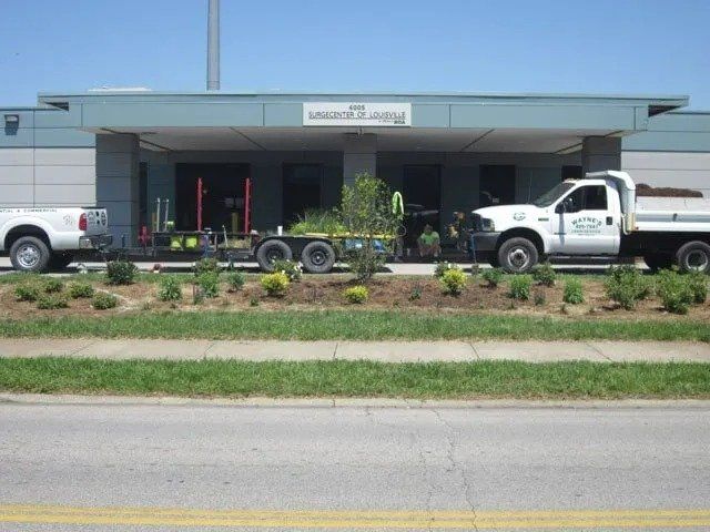 Landscapers with trucks and trailer planting bushes in front of a building with a sign that reads 