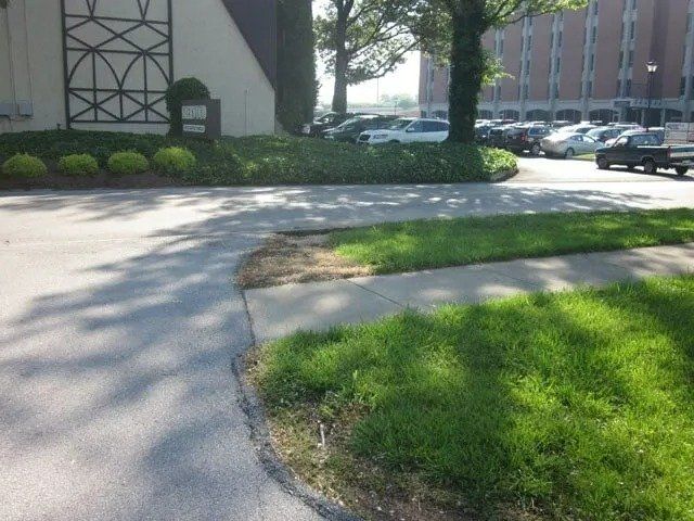 A paved driveway curves past a patch of green grass and landscaping near a building, with parked cars in the background.