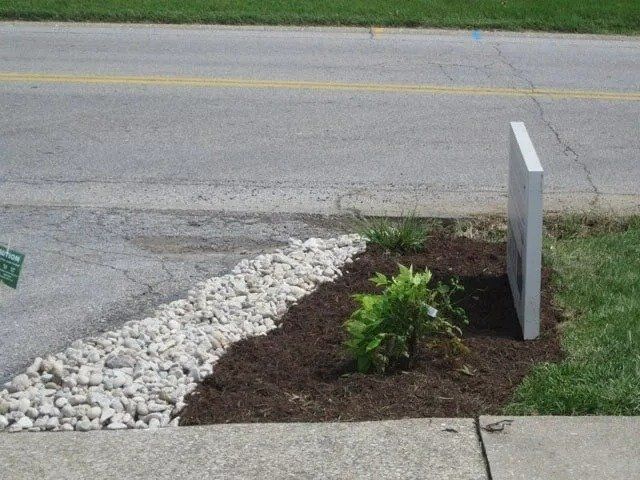 A landscaped area with white rocks, brown mulch, and a small plant next to a sidewalk and street with a sign.