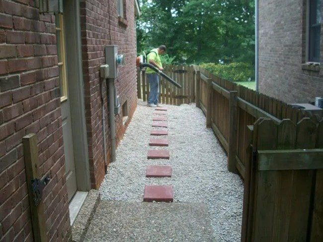 Person using a leaf blower on a gravel path with stepping stones between two brick buildings and a wooden fence.