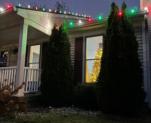 House exterior at dusk, decorated with Christmas lights, with a lit Christmas tree visible inside a window. House exterior at dusk, decorated with Christmas lights, with a lit Christmas tree visible inside a window.