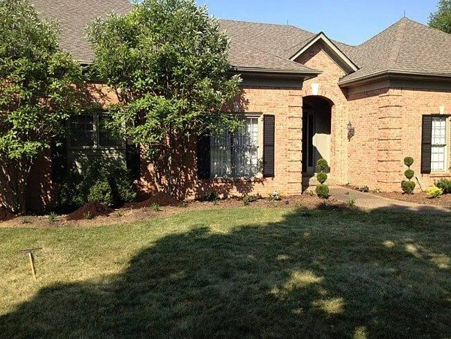 A brick house with a brown roof, green lawn, and landscaping. Trees cast shadows across the yard.