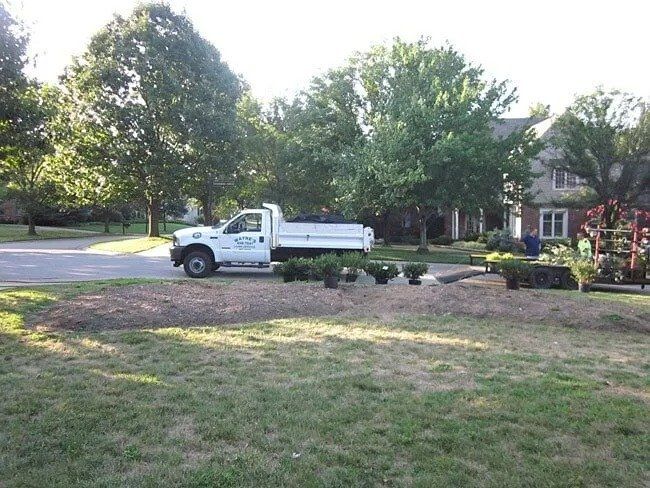 A white landscaping truck parked on a residential street, next to some shrubs and a prepared garden bed.