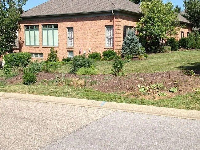 Red brick house with manicured lawn and flower beds beside a paved road; mature trees in the background.