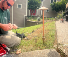 A man installing a landscape light in a residential yard; grass, house, and a walkway are visible. A man installing a landscape light in a residential yard; grass, house, and a walkway are visible.