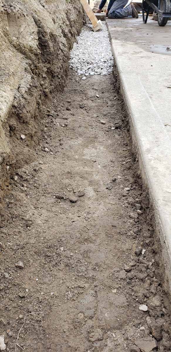 A person is digging a trench in the ground next to a sidewalk.