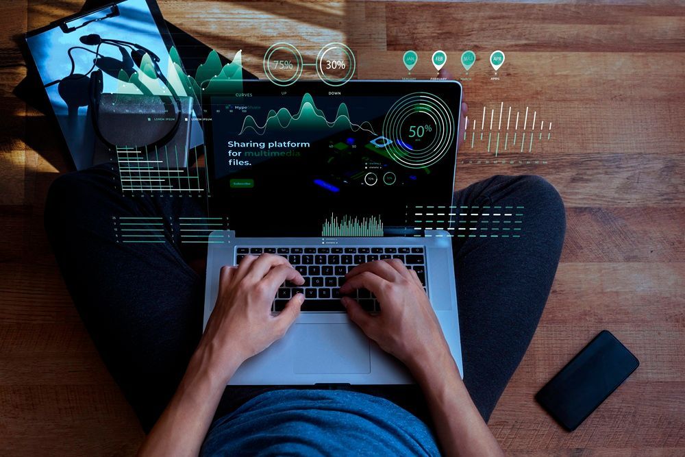 Person typing on laptop, overlaid with data visualizations, sitting on wooden floor next to a phone.