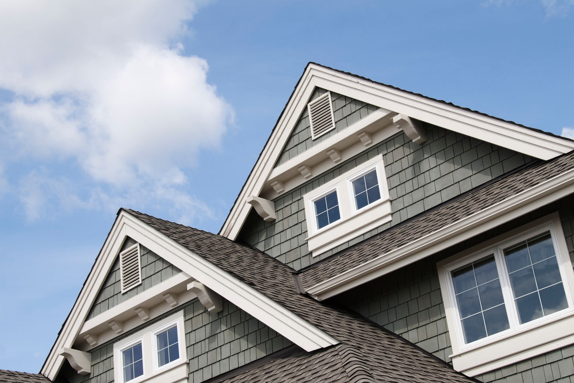 Close-up of a house exterior with dark roof, blue-green siding, white trim, and a blue sky with clouds.