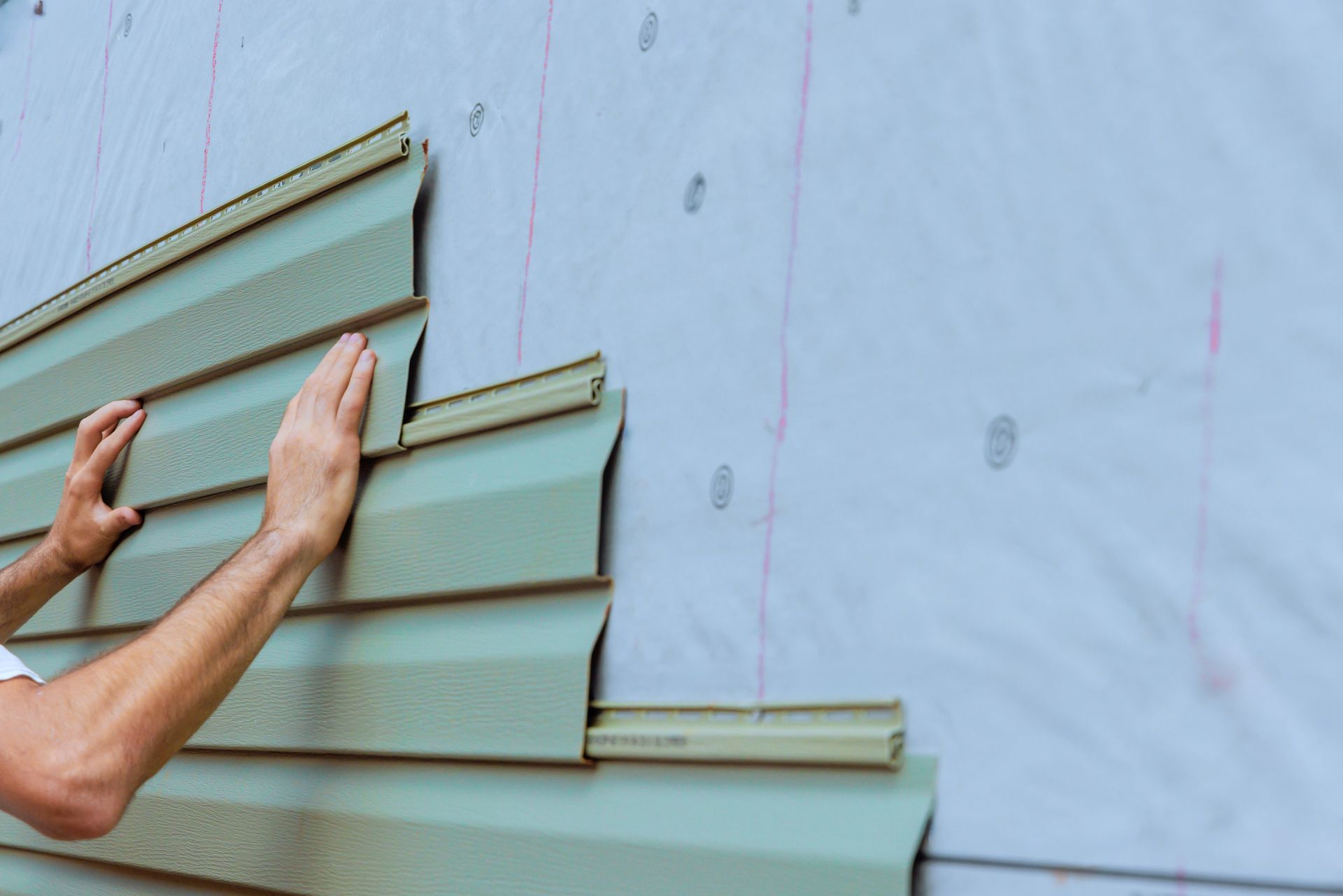 Hands installing light green vinyl siding on a house exterior covered in white protective material.