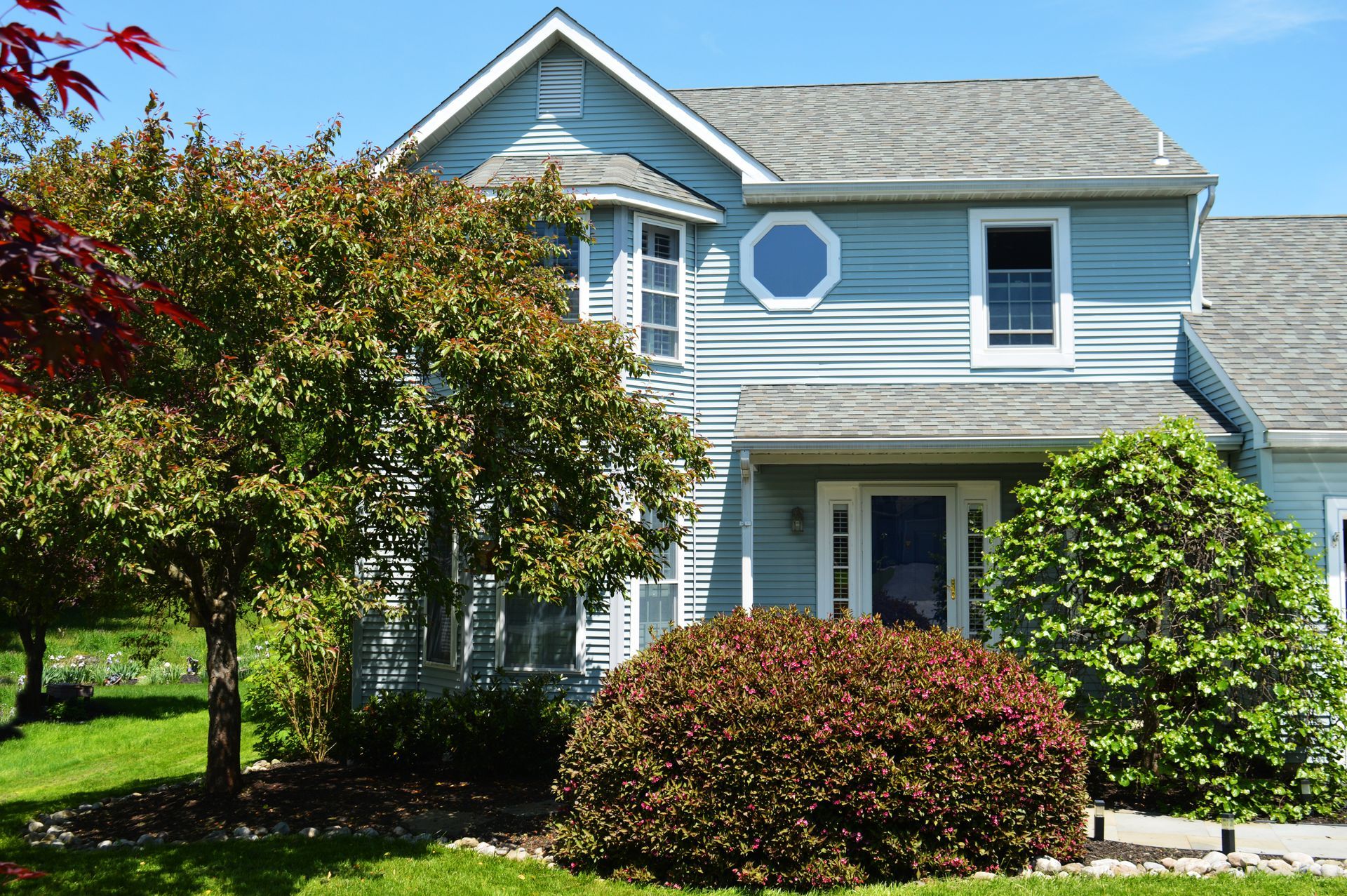 Blue house with bay window and shrubs in front. Green lawn and trees.