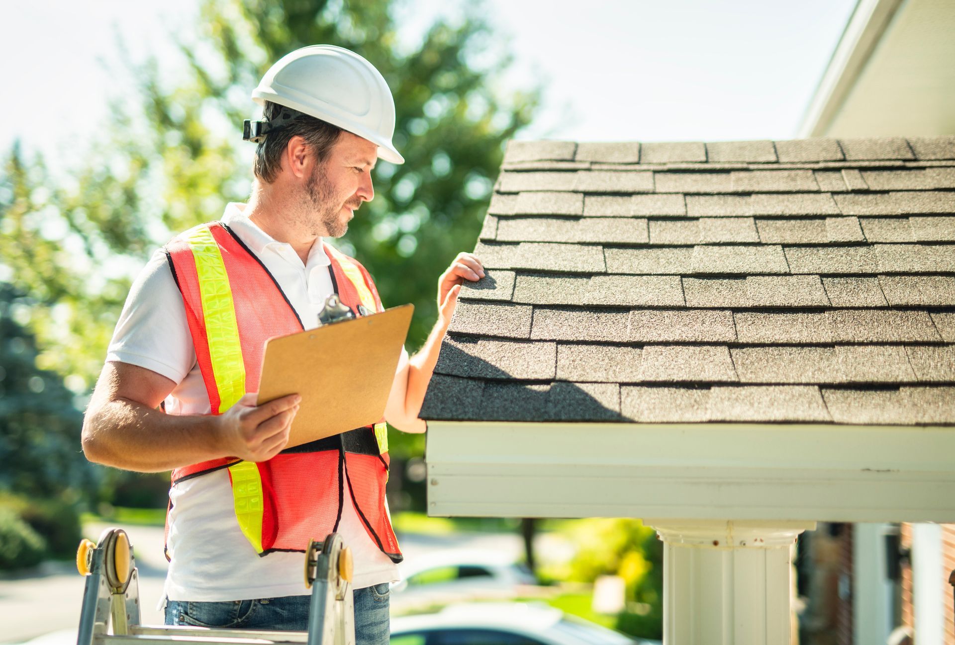 Man in safety vest and hard hat inspecting a roof, holding a clipboard outdoors.