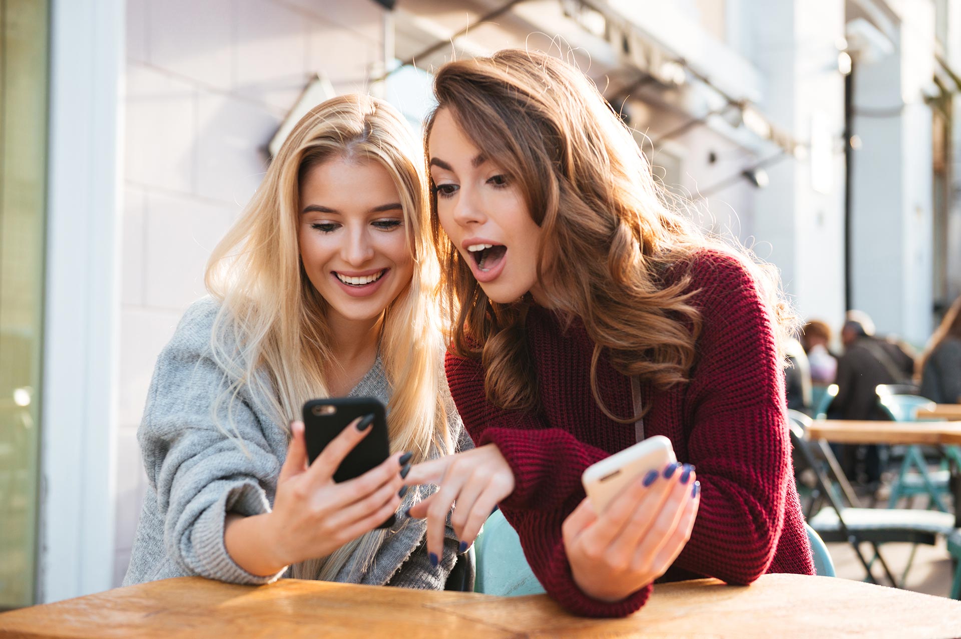 Two excited young girls using mobile phones while sitting at the cafe outdoors and pointing finger