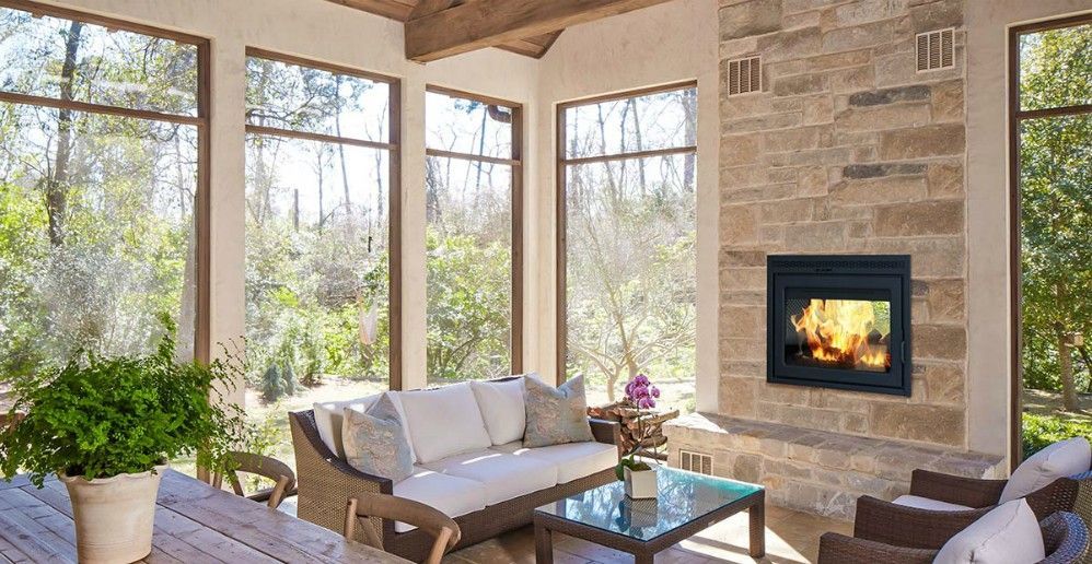 Sunroom with stone fireplace, glass windows, beige sofa, and surrounding greenery.