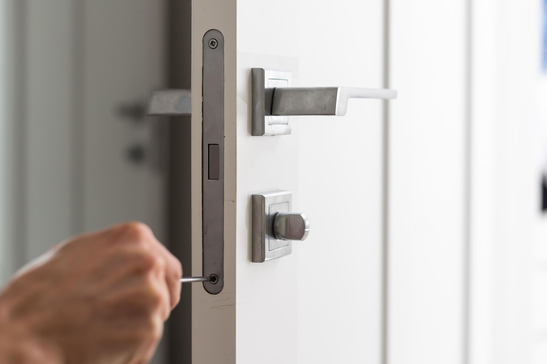 Hand using a screwdriver to tighten a screw on a door latch plate; white door, metal hardware.