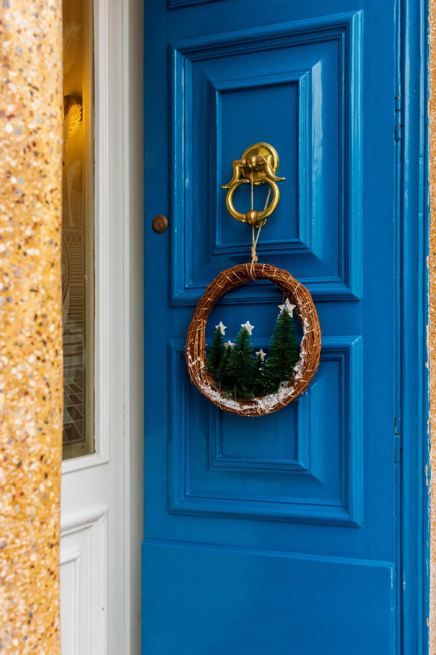 Blue door with a gold knocker and Christmas wreath, next to a white door and textured wall.