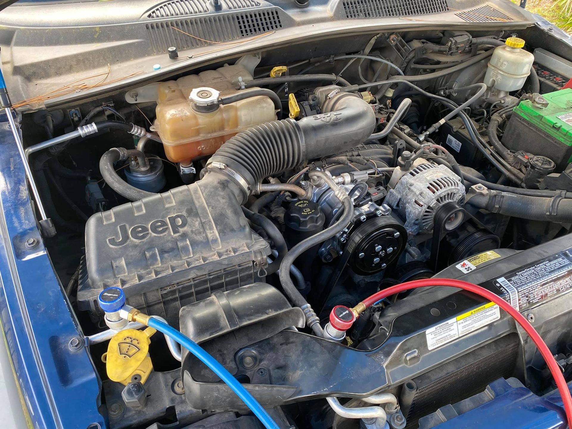 Engine bay of a blue Jeep with AC service gauges attached. Black air filter box labeled 