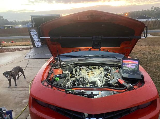 Orange car with open hood, laptop, tools, and a dog. Outdoors, near a trailer.
