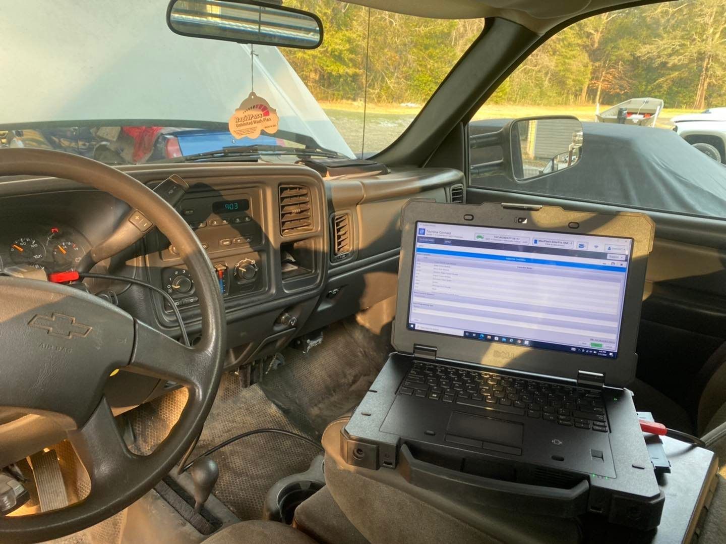 Laptop on a car seat displays diagnostic software. Dashboard, steering wheel, and open hood visible.