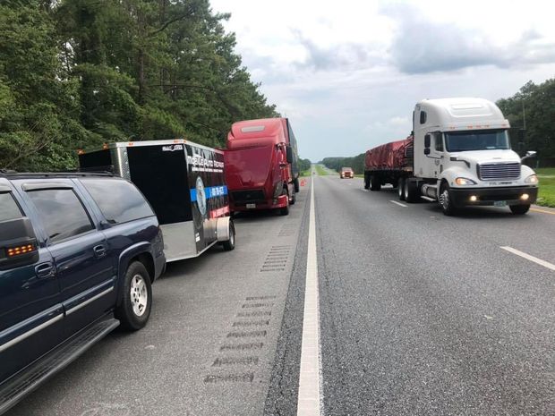 Vehicles stopped on a highway shoulder. Includes a truck with trailer, a red semi-truck, and a white semi-truck with cargo.