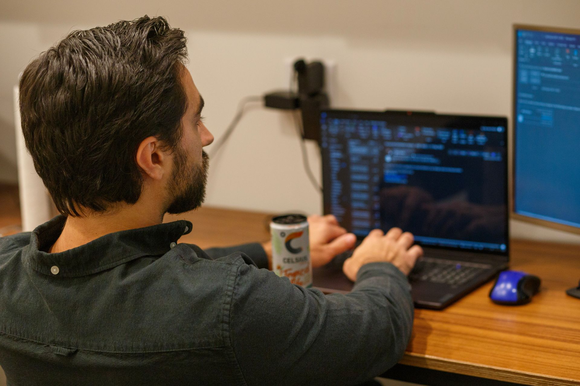 Man with dark hair and beard, coding on a laptop with a second monitor, drink on desk.