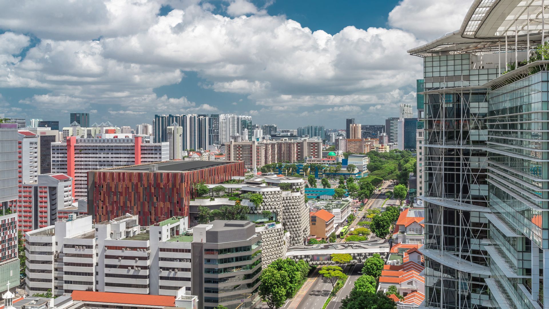 A wide cityscape view of modern buildings with varied architectural styles, greenery, and a cloudy blue sky.