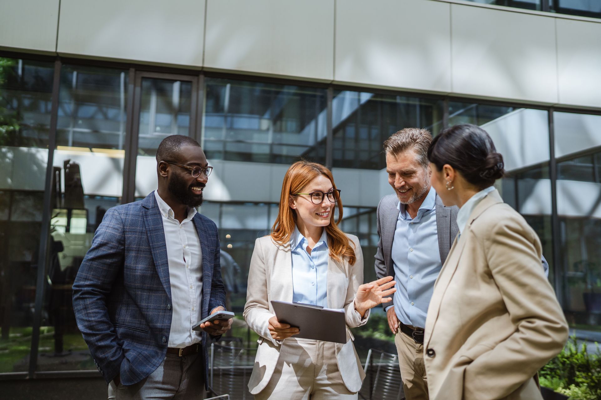 Four coworkers talking outside an office building, with one holding a tablet and another smiling in a beige blazer