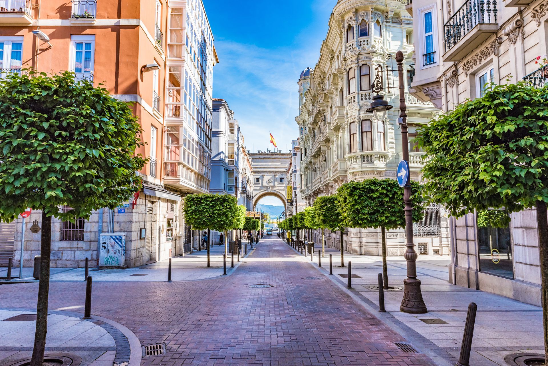Empty cobblestone street lined with trees and ornate buildings, framing an archway in the distance