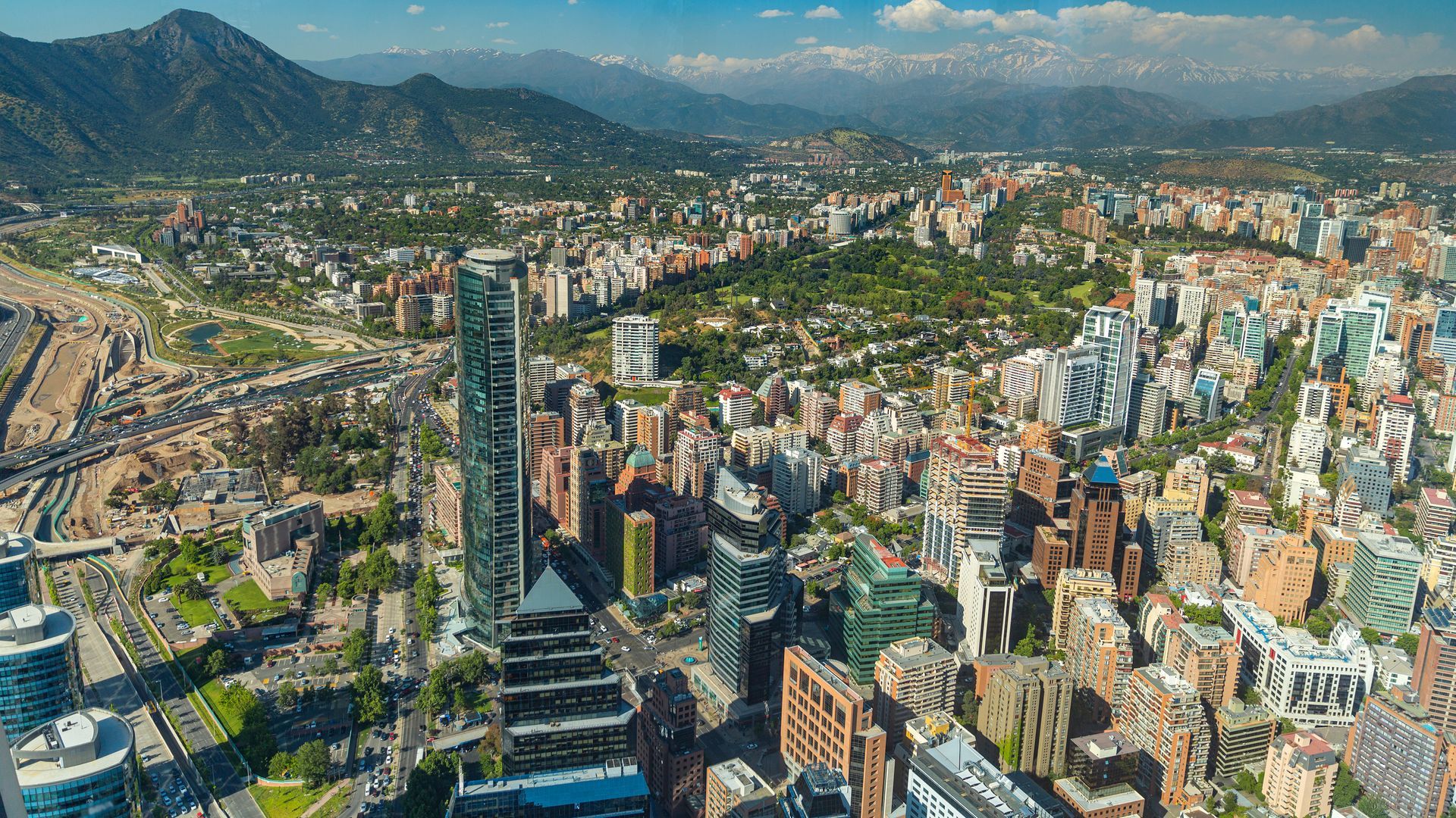 Aerial view of a dense city skyline with tall towers, mountains in the background, and a broad highway on the left.