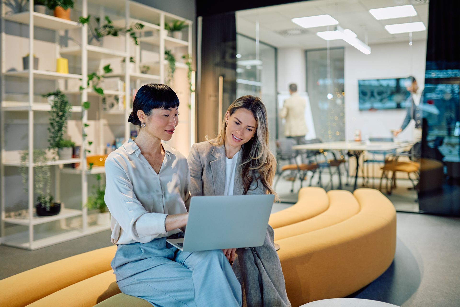 Two professionals sit on a curved yellow sofa in a modern office, collaborating while looking at a laptop screen.