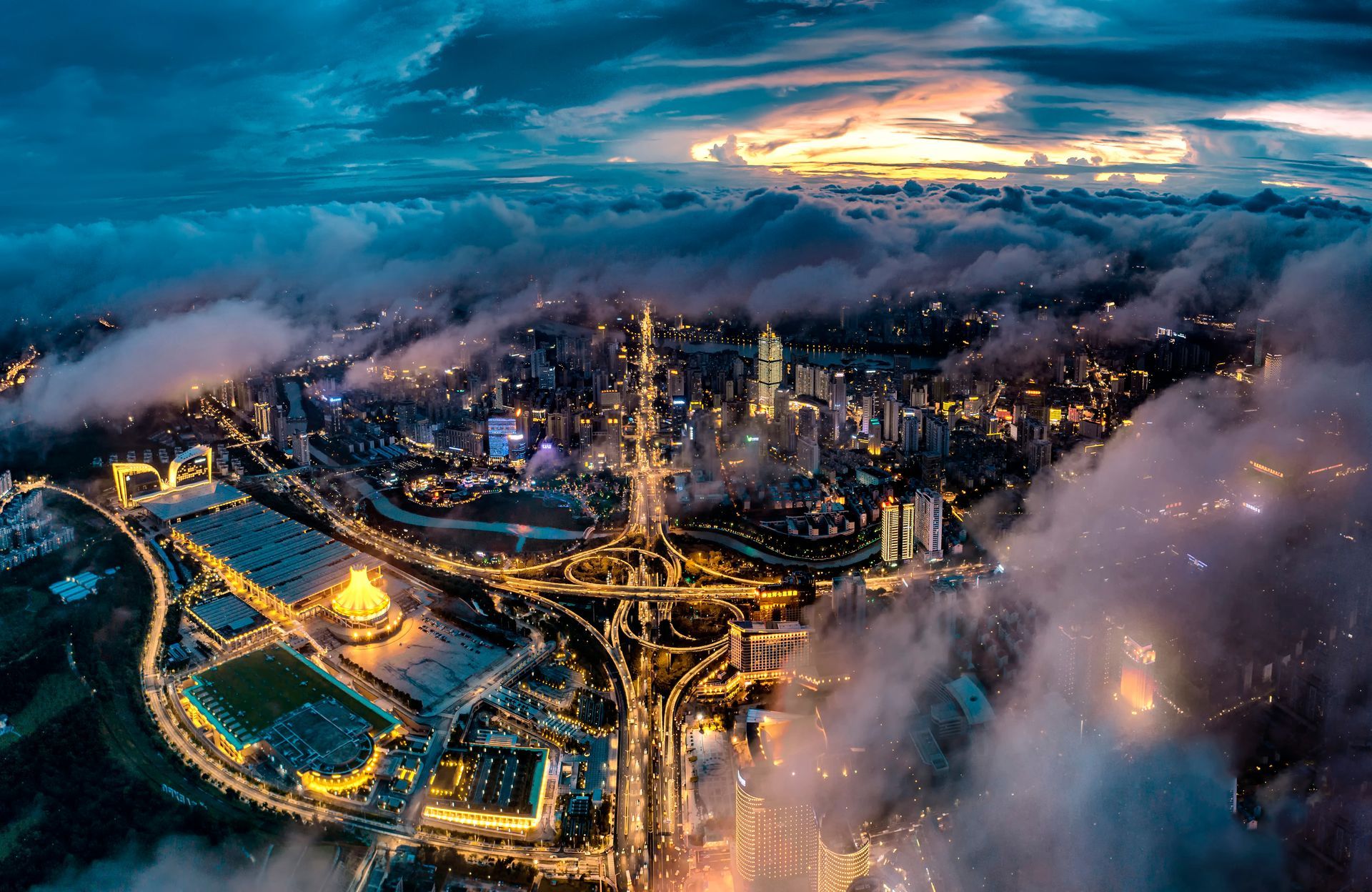 Aerial view of a city at dusk with glowing highways and skyscrapers under low clouds