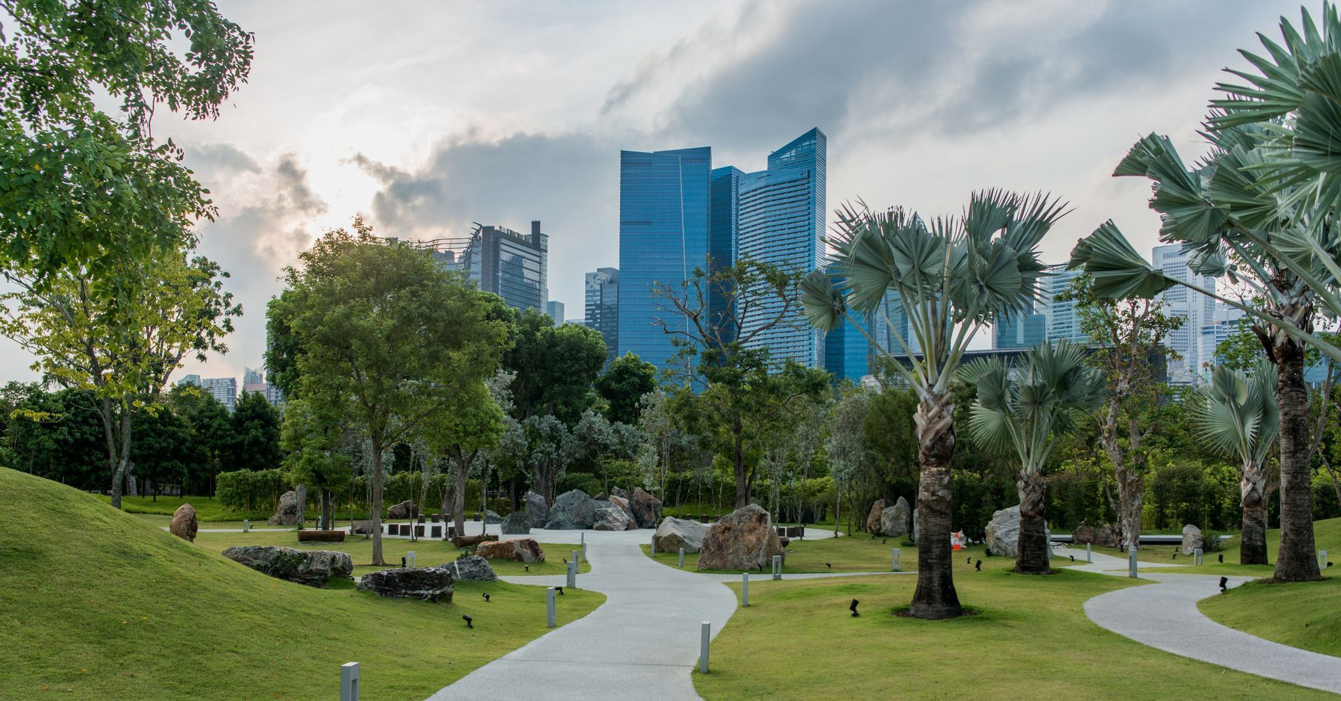 Park with winding pathways, trees, and city skyline in the background under a cloudy sky.