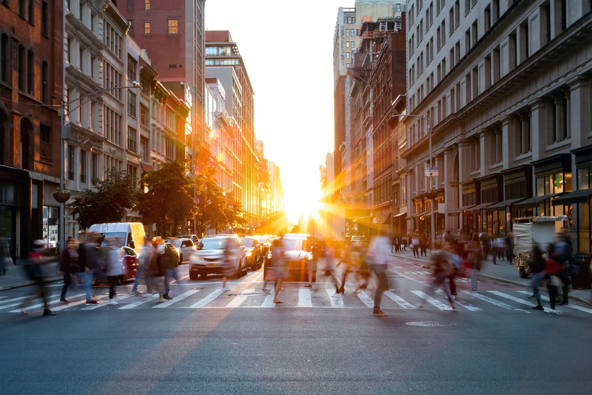 City street with pedestrians crossing; sun shining between buildings, golden light.