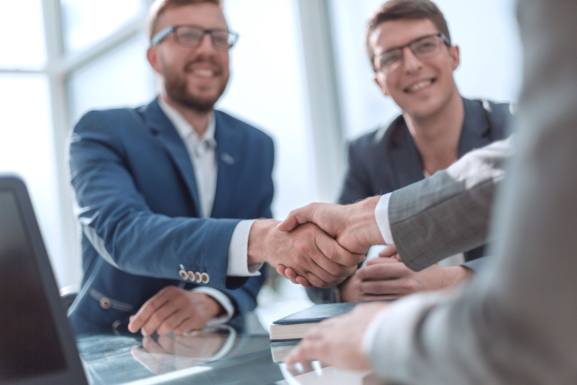Three men in suits shake hands, smiling, at a glass table in an office setting.