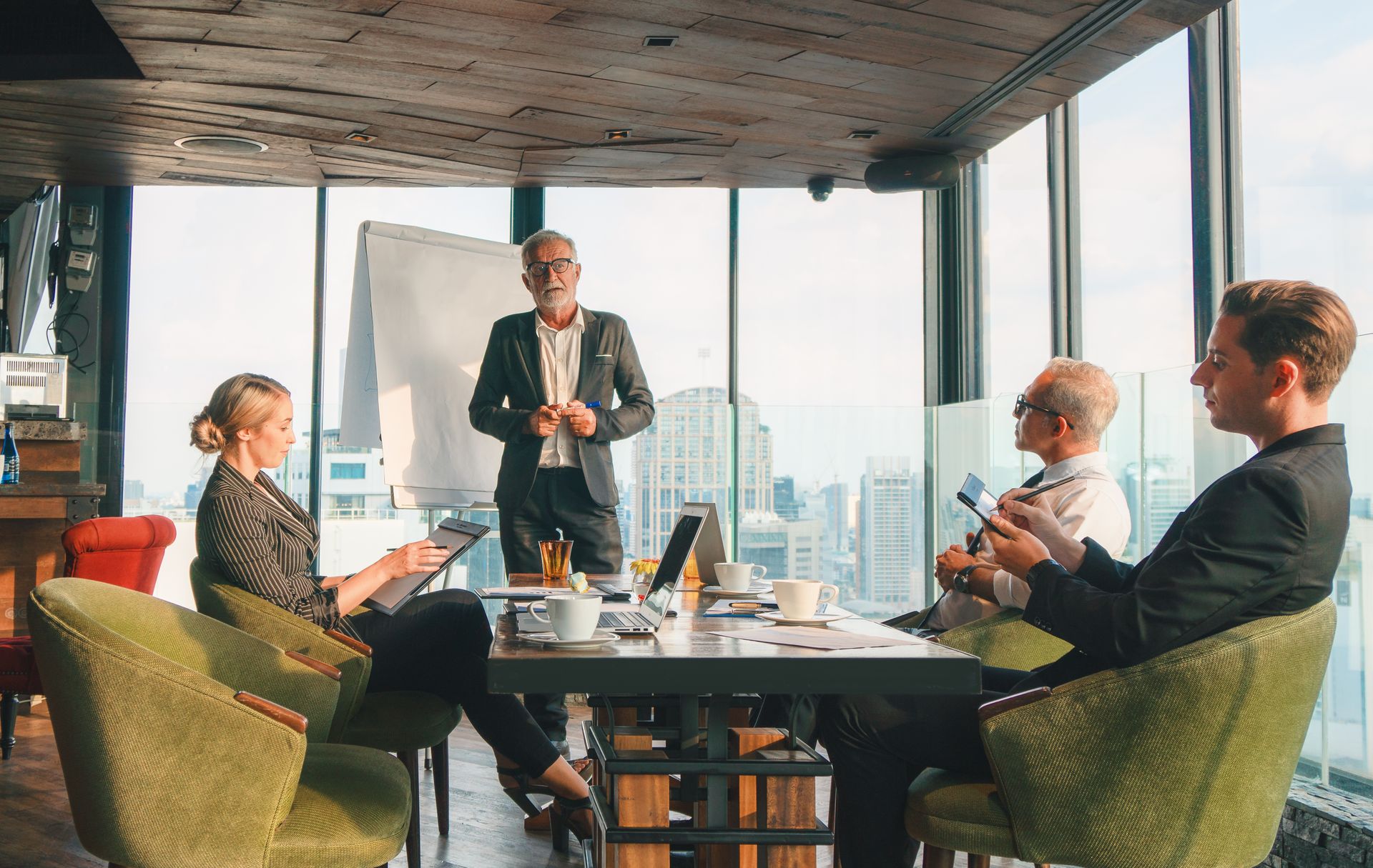 A group of professionals in suits meets in a high-rise conference room with a city view while a colleague presents.