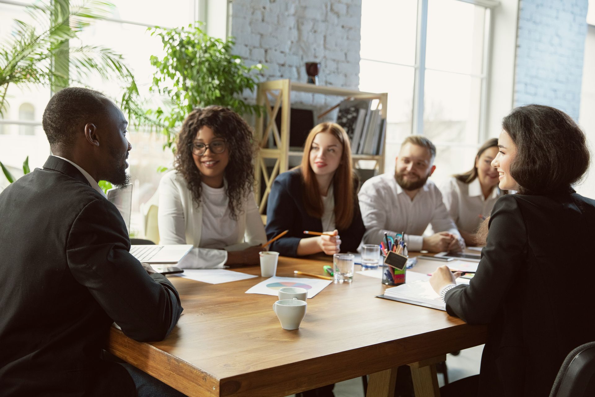 People in a bright meeting room discussing around a wooden table with notebooks and cups
