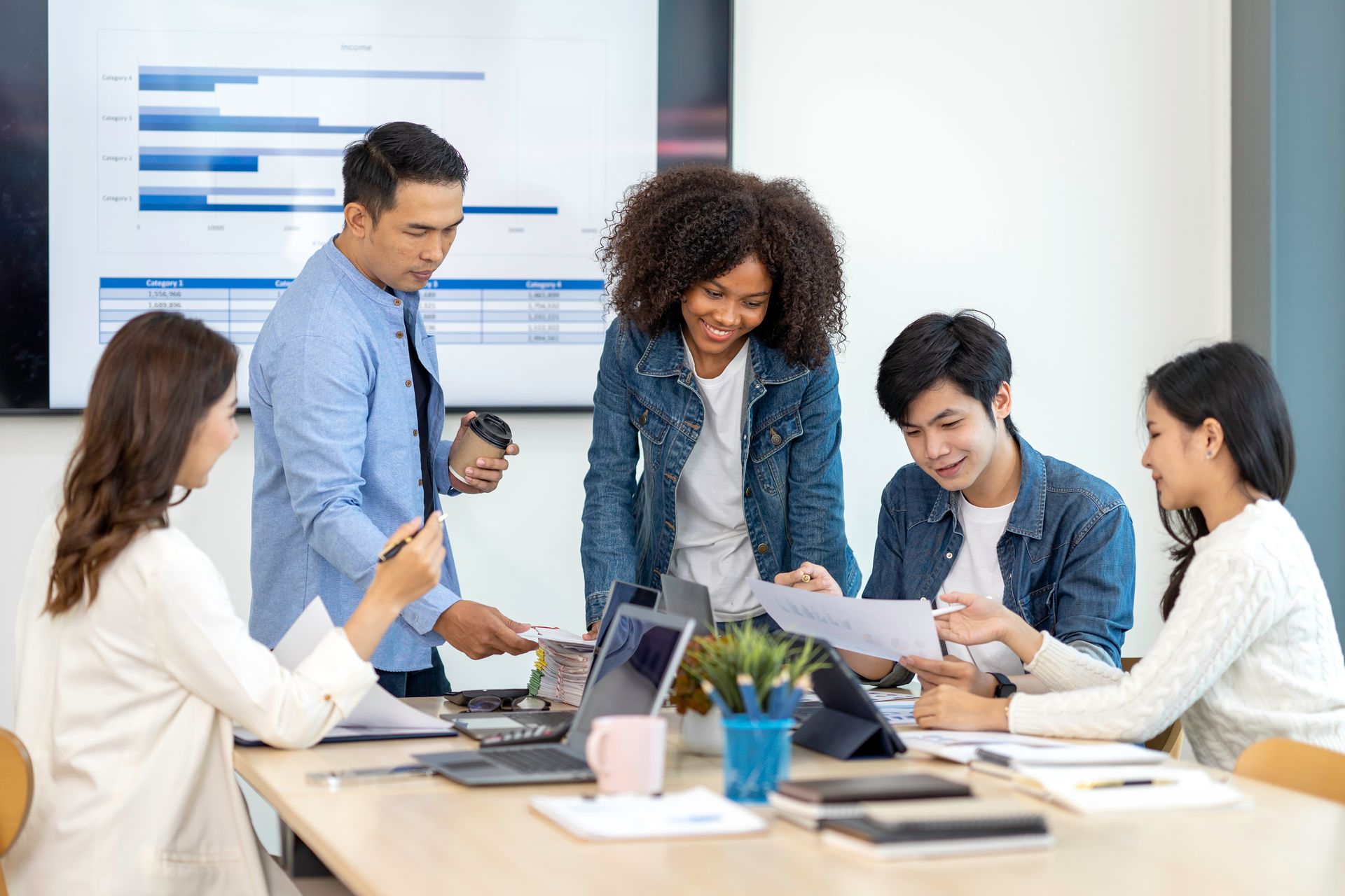 Coworkers in a bright office review papers and discuss a project around a table with laptops and notebooks