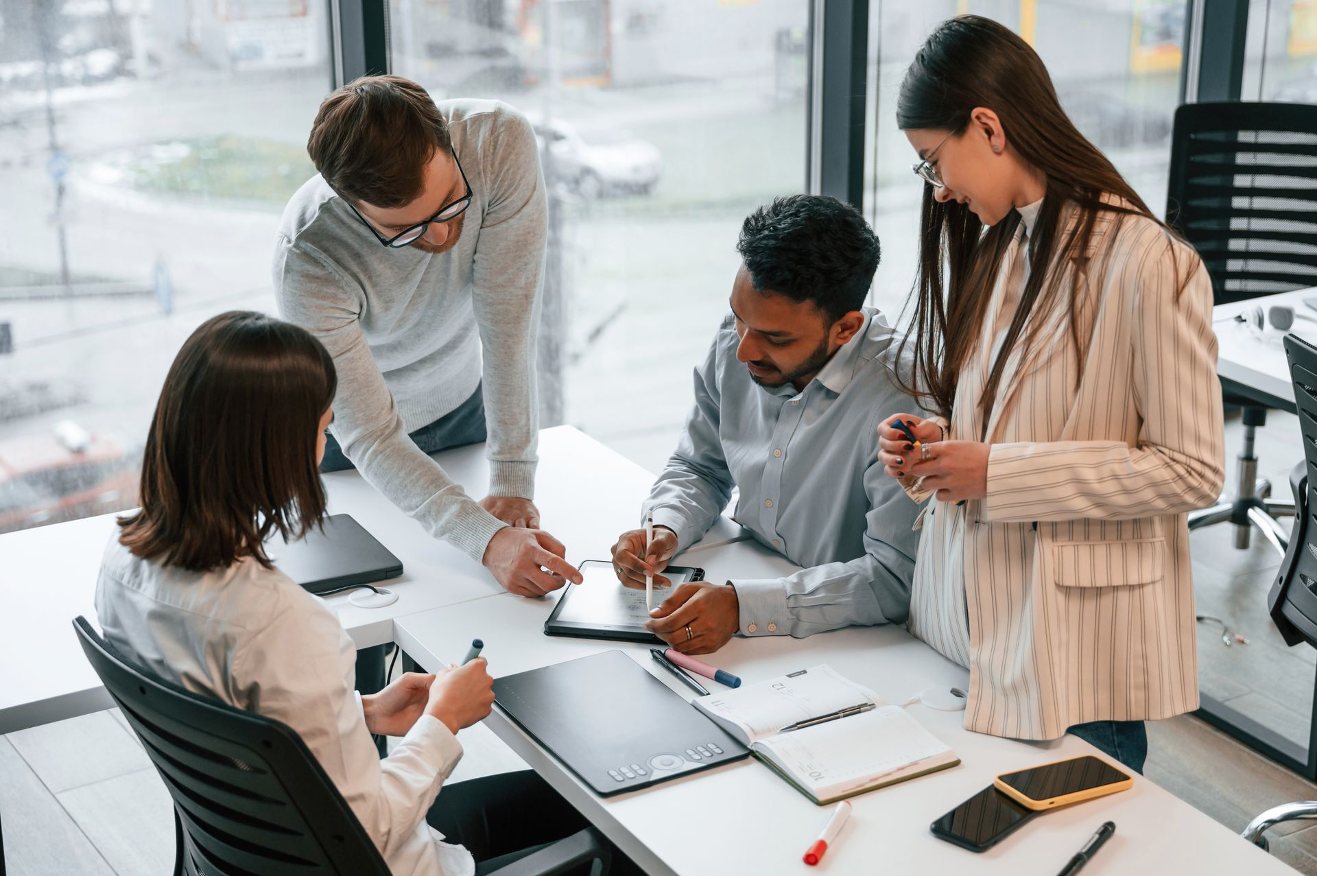 Four people in an office, discussing documents at a table. Sunlight streams through a large window.